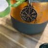 Vegvisir Pendant Amulet Necklace The Vegvisir Pendant Amulet Necklace, featuring Norse runic designs, hangs on a string in front of a green potted plant with an orange and gray pot, resting on wood with polished stones nearby.