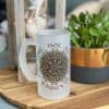 A Vegvisir Nor all who wander are lost frosted glass mug sits on a wooden tray next to a potted plant and a decorative glass container of seashells.