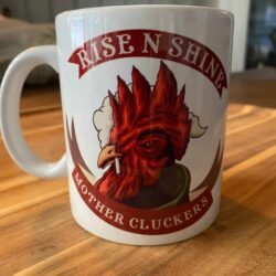 The Rise n Shine Mother Cluckers Mug is a white coffee mug with a grumpy red rooster smoking a cigarette, RISE N SHINE above and MOTHER CLUCKERS below in bold letters, resting on a wooden surface.