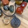 A Valknut Pendant Amulet Necklace with a Celtic knot design rests on a wooden surface, displayed on a black cord and surrounded by polished multicolored stones in red, green, yellow, and gray.