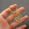 A hand holds a Custom Name Tree Of Life Stainless Steel Keychain, featuring a round gold design with a tree and the names Paco, Margaret, Sheila, and Ismael engraved in cursive on its branches.