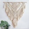 The Nordic Bohemian Macrame Wall Hanging displays detailed knots and tassels in a triangular design on a wooden dowel, with a green leafy plant in the lower left corner enhancing its boho decor charm.
