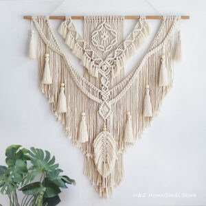 The Nordic Bohemian Macrame Wall Hanging displays detailed knots and tassels in a triangular design on a wooden dowel, with a green leafy plant in the lower left corner enhancing its boho decor charm.