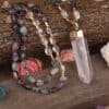 Natural White Quartz Stick Point Pendant Rainbow Fluorite Stone Chip 108 Prayer Beads Knotted Handmade Necklace N0280AMCD Close-up of the Natural White Quartz Stick Point Pendant Rainbow Fluorite Stone Chip 108 Prayer Beads Knotted Handmade Necklace N0280AMCD on a wooden surface, surrounded by decorative stones and artificial flowers.