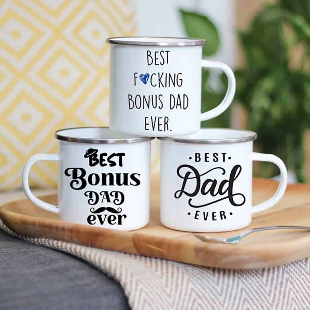 Set of three Dad, Step Dad Bonus Dad Enamel Mug white mugs with metal rims on a wooden tray, each with a unique Best Bonus Dad Ever message in black font. Perfect for any step dad. Cozy scene features a yellow patterned pillow.
