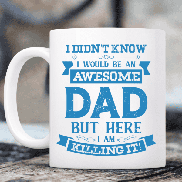 I'M An Awesome Dad Coffee Mug The IM An Awesome Dad Coffee Mug, white with blue text that reads “I didn’t know I would be an awesome dad but here I am killing it!”, rests on a table with a softly blurred background.