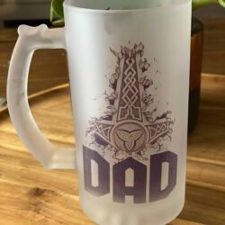 The Viking Thor’s Hammer Custom Beer Mug features a frosted glass design with a bold Thor’s Hammer above “DAD,” displayed on a wooden surface with a green plant in the background.