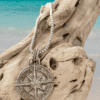 The Men’s Compass Necklace features a silver pendant resting on weathered driftwood, with turquoise ocean water and a clear blue sky in the background.