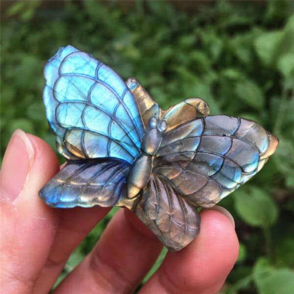 A hand holds a Natural Blue Labradorite hand carved butterfly with iridescent blue and brown wings, detailed carving, and a blurred green foliage background.