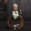 Little Buddha Backflow Incense Burner The Little Buddha Backflow Incense Burner features a small ceramic Buddha in green robes atop a wooden ring, with an incense cone beside it releasing smoke that gently cascades down through the ring.
