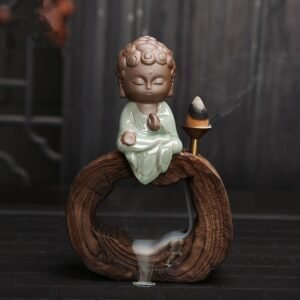 The Little Buddha Backflow Incense Burner features a small ceramic Buddha in green robes atop a wooden ring, with an incense cone beside it releasing smoke that gently cascades down through the ring.