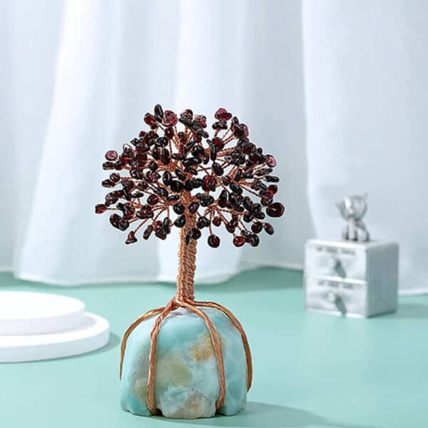 Natural Gem Stone & Copper Feng Shui Trees A Natural Gem Stone & Copper Feng Shui Tree with wire branches and red gemstone leaves on a blue stone base is displayed on a light green surface, with a white curtain and small silver ornament in the background.