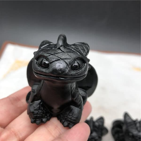 A hand holds a small, black Obsidian Toothless Dragon figurine with large eyes and detailed scales, inspired by How to Train Your Dragon. The background is blurred.