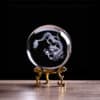 The Chinese Dragon Crystal Ball with Stand showcases an intricate dragon design inside a clear sphere, displayed on an ornate gold stand atop a wooden surface against a black backdrop.
