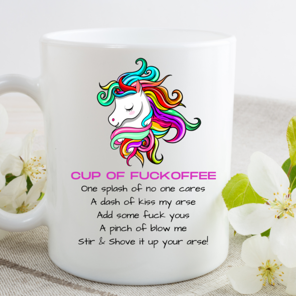 The Cup of Fuckoffee! Coffee Mug, featuring a colorful unicorn and a funny recipe starting CUP OF FUCKOFFEE, sits on a white table beside blooming white flowers and green leaves.