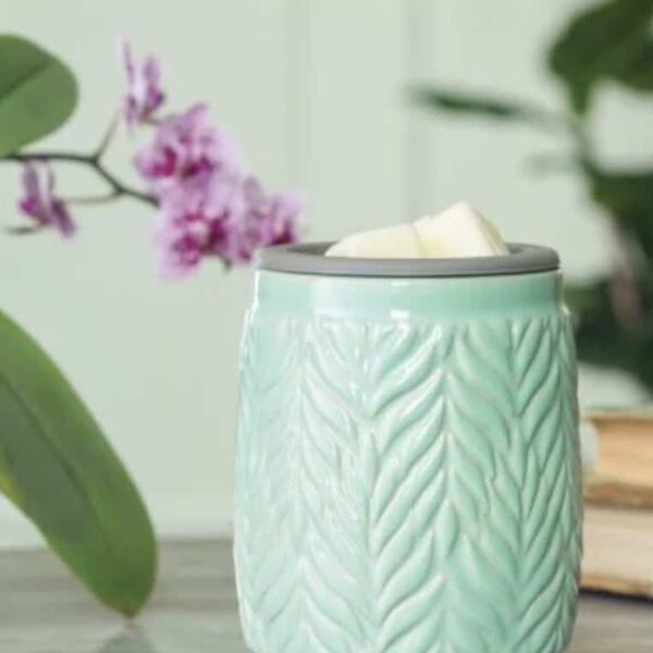 Mint Flip Dish Wax Warmer The Mint Flip Dish Wax Warmer, featuring a leaf pattern, holds white wax melts on a table. A softly blurred purple flower and green leaves appear in the background.