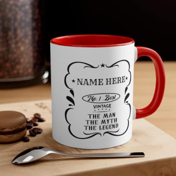 A Personalised The Man Myth Legend Mug with a red handle rests on wood beside a spoon, macaron, and coffee beans. Its text reads: Name Here. No. 1 Best Vintage. The Man. The Myth. The Legend—an ideal custom gift!.