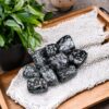 A wooden tray displays polished Snowflake Obsidian Tumble Stones on a woven cloth, accented by green potted plants.