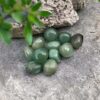 A cluster of Aventurine Tumble Stones rests on a rough gray rock, with green leaves and a textured stone in the background.