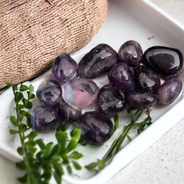 Yellow Jasper Tumble Stone A white tray displays several small Amethyst Tumble Stones, complemented by green leafy sprigs and a hint of textured pink fabric in the background.