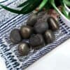 A small pile of shiny, dark Pyrite Tumble Stones rests on patterned blue and white fabric, with green leaves from a potted plant visible in the background.