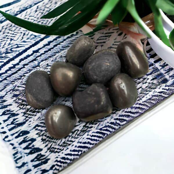 Yellow Jasper Tumble Stone A small pile of shiny, dark Pyrite Tumble Stones rests on patterned blue and white fabric, with green leaves from a potted plant visible in the background.