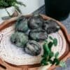 Emerald Tumble Stone Crystal A wooden tray displays six polished Emerald Tumble Stone Crystals, their rich green and black hues gleaming atop a woven mat, surrounded by lush potted plants and greenery.