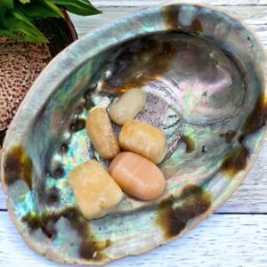 A Yellow Calcite Tumble Stone, smooth and polished, sits inside an iridescent abalone shell on a white wooden surface, with part of a plant in the background.