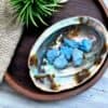 Blue Magnesite Tumble Stone Several Blue Magnesite Tumble Stones are displayed in an iridescent abalone shell on a round wooden tray, accompanied by a green artificial air plant and beige textured fabric on a light-toned surface.
