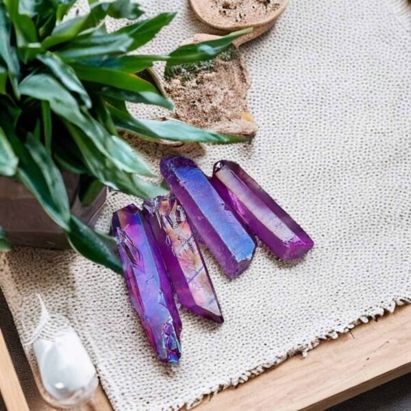Four Purple Aura Angel Quartz Points rest on a textured beige mat atop a wooden tray, surrounded by green plants and a small sand dish accented with greenery.