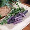 A Lepidolite Mini Tower Crystal, polished to a shine, sits with other purple stones on a burlap cloth accented by green leaves. In the background, a woven basket with a leafy plant rests on a wooden surface.