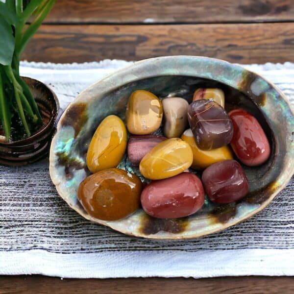 White Howlite Tumble Stone A ceramic bowl filled with Mookaite Tumble Stones in yellow, red, brown, and tan rests on a woven cloth beside a small potted plant atop a wooden table.
