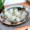 A large, iridescent shell holds smooth, polished New Jade Tumble Stones and rests on a woven mat beside a potted plant atop a wooden surface.