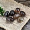 Six polished, oval Smokey Quartz Tumble Stones are arranged on a textured beige mat with a green leafy plant in the corner, all set on a dark wooden surface.