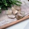 Six smooth Rutilated Quartz Tumble Stones with light brown and golden hues are arranged on a textured beige cloth, framed by a wooden tray with green plant leaves in the background.