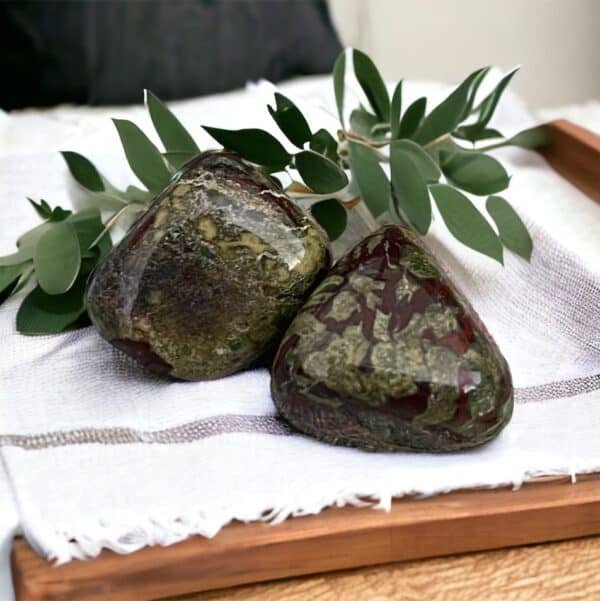 White Howlite Tumble Stone Two polished Dragon Blood Jasper Tumble Stones, featuring green with red and brown hues, rest on a white cloth with a leafy branch behind them, displayed on a wooden tray.
