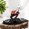 Engraved Decanter and Glass Set The Engraved Decanter and Glass Set features a modern decanter on a black stand, paired with two matching glasses, all displayed on a rustic wooden surface beside a green plant.