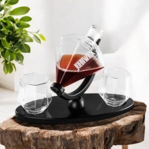 The Engraved Decanter and Glass Set features a modern decanter on a black stand, paired with two matching glasses, all displayed on a rustic wooden surface beside a green plant.