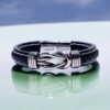 Men’s Personalised Link Bracelet The Men’s Personalised Link Bracelet features black braided leather, a silver clasp with a knot design, and is engraved with Linked Forever. Its showcased on a glossy pale surface with a softly blurred blue and purple background.