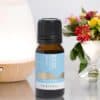 A small amber bottle labeled Pisces Zodiac Blend rests on a white surface, with a diffuser and a vase of colorful flowers softly blurred in the background.