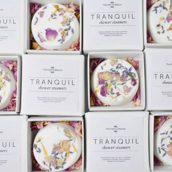 Open white boxes of Tranquil Individual Shower Steamers, each topped with pink and yellow dried flower petals. Arranged in a grid, the packaging reveals both the shower steamer and its product label inside.