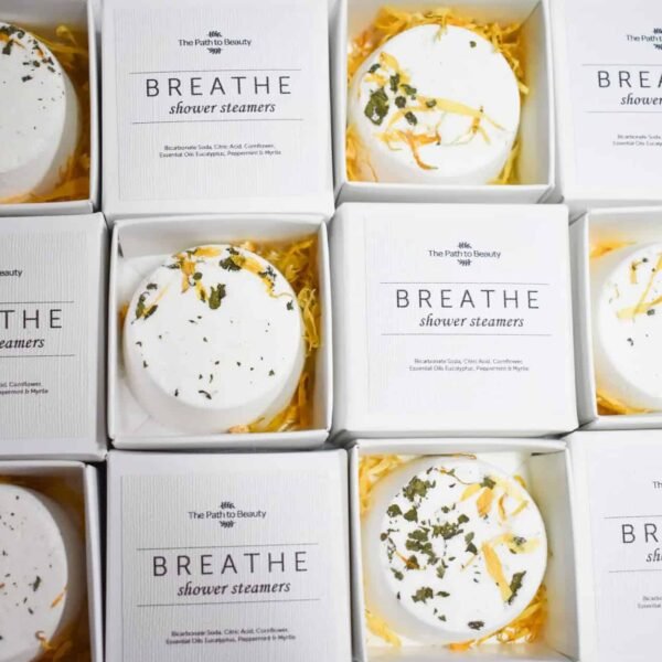 Breathe Individual Shower Steamers Nine boxes of Breathe Individual Shower Steamers are arranged in a grid. Each round, white steamer with dried herbs rests in yellow shredded paper inside its labeled box.