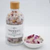 A clear glass bottle labeled Tranquil Bath Salts holds colorful salts, sitting beside a small white bowl with a portion of the mixture, featuring visible flower petals and crystals for ultimate relaxation.