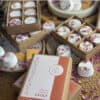 Geranium Lavender Shower Steamers Boxes of round, white Geranium Lavender Shower Steamers, infused with floral scents and topped with dried petals, are scattered on a woven surface. One box is labeled Botanicals Shower Steamer Vault.
