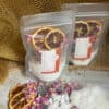 Two pouches of Lavender & Rose Geranium Botanicals Coconut Bath Milk with dried orange slices and flower petals are displayed on a textured surface. Bath milk, petals, and orange slices are spread in front; gold mesh decor is in the background.