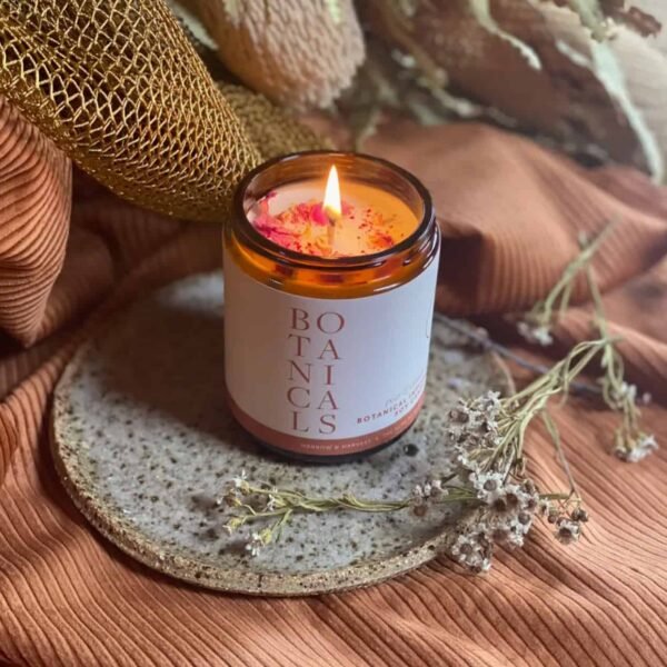 Pear & Cinnamon Botanical Candle The Pear & Cinnamon Botanical Candle in an amber glass jar sits on a speckled ceramic plate, surrounded by dried flowers and textured brown fabric, creating a warm and cozy ambiance.