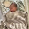 Custom Personalised & Photo Blankets A newborn sleeps peacefully in a cozy beige and white Custom Personalised & Photo Blanket with the name Otto. A round wooden sign says Hello, Im new here, set against a soft background of green leafy fabric.