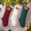 Three Personalised Knitted Christmas Stockings—red for Jackson, white for Olivia, and green for Liam—hang under festive pine branches, each featuring a name tag.