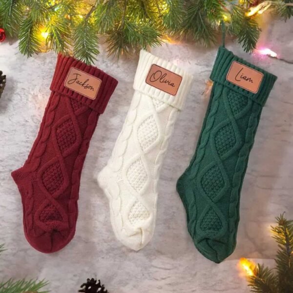 Three Personalised Knitted Christmas Stockings—red for Jackson, white for Olivia, and green for Liam—hang under festive pine branches, each featuring a name tag.