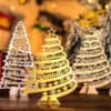 Three Personalised Christmas Tree ornaments in Gold, Silver, or Wood, each with custom names and the year 2021, arranged on a festive table with blurred lights and mini trees in the background.